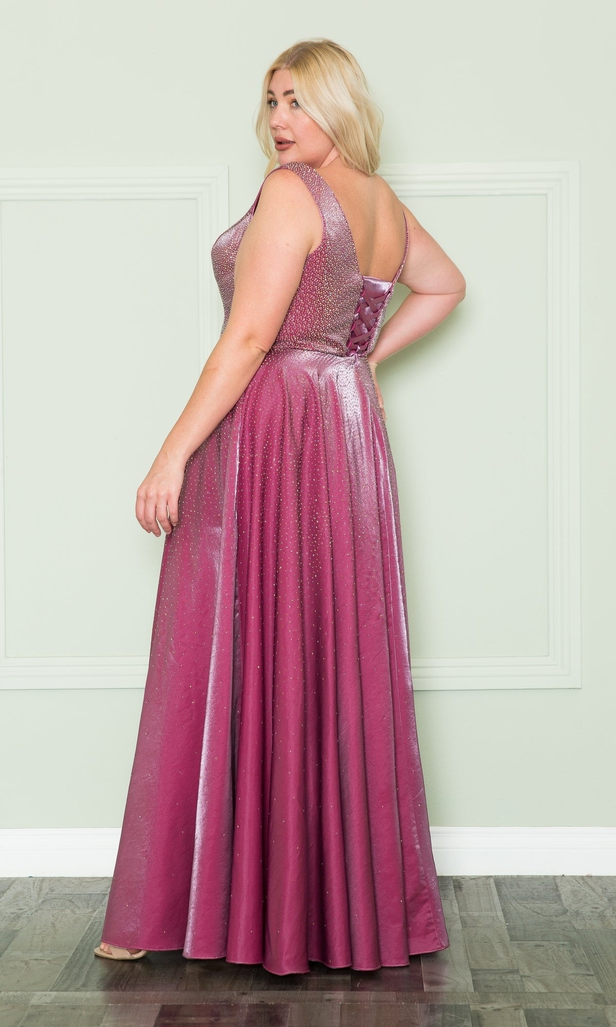 A woman with blonde hair stands against a light wall and wood floor in the Embellished Plus-Size Long V-Neck Prom Dress W1110—a sleeveless, shimmery, magenta gown with a fitted bodice and full skirt.
